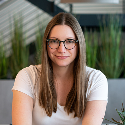 Porträt von Lara aus dem Team Marketing. Sie sitzt an einem Tisch, hat glatte, lange Haare, trägt eine Brille und ein weißes T-Shirt und lächelt leicht in die Kamera.